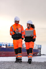 Two engineers wearing high-visibility safety gear and hard hats review plans on a laptop at a...