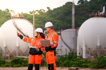 Two engineers in safety gear review project plans on a laptop and use a walkie-talkie at an...