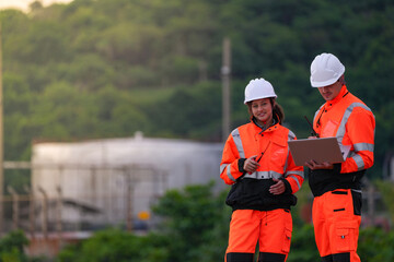 Two engineers in safety gear review project plans on a laptop and use a walkie-talkie at an...