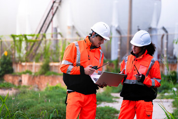 Two diverse engineers in safety gear collaborate with a laptop at a natural gas storage facility. A...