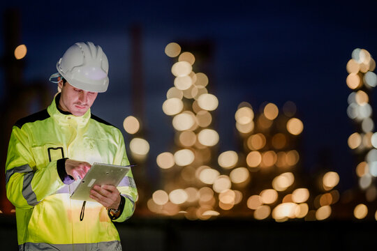 A dedicated engineer in a hard hat uses a digital tablet during a night shift at a refinery. A concept for modern industry, on-site data analysis, and 24-hour operations.