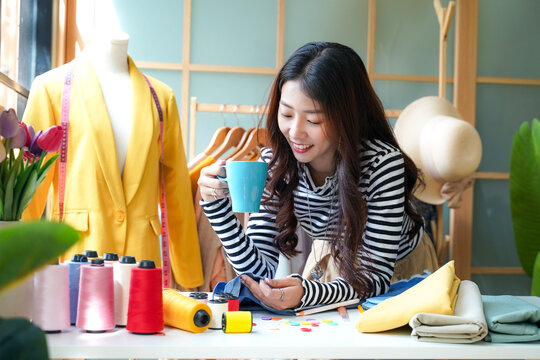 Asian young women fashion designer drinking coffee relax after working on her designer in the showroom, Lifestyle Stylish tailor use computer clothing design in studio. Business small Concept