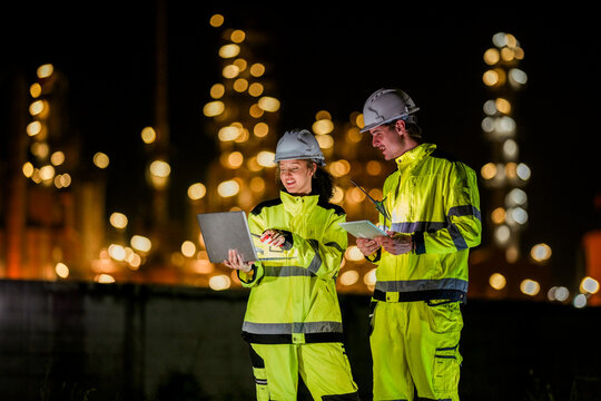 Two diverse engineers coordinate operations on a night shift. The man uses a radio while the woman explains data from a digital tablet, a concept for industrial communication.
