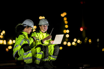 Two industrial workers in high-visibility gear coordinate using radios and a laptop at night, with...