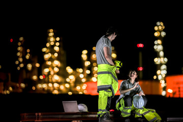Two industrial workers take a break during the night shift at a construction site, sitting on steel...