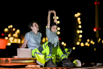 Two industrial workers take a break during the night shift at a construction site, sitting on steel...