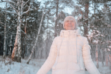 joyful woman stands in snowy forest, hands raised, as snowflakes fall around her. she wears warm...