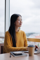 Young Asian businesswoman working on laptop and looking out the window, taking a break