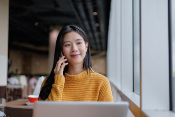 Young Asian Freelancer Woman Talking on Phone and Working on Laptop in Cafe