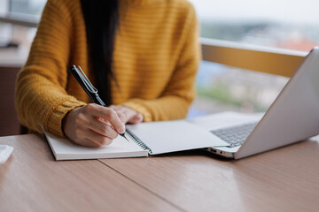 Woman writing notes on notebook while working on laptop computer