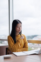 Young Asian businesswoman working and thinking in a modern office with laptop and notebook