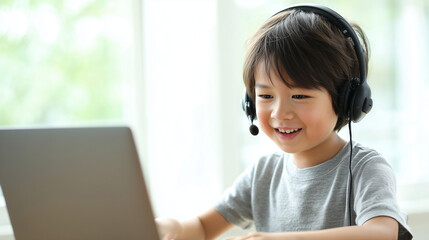 明るい室内で、ヘッドセットをつけた小学生くらいの日本人の男の子が、笑顔でノートパソコンを操作している様子。背景は窓からの光で白くぼけており、清潔感がある。オンライン授業や学習をイメージさせる、親しみやすい雰囲気
