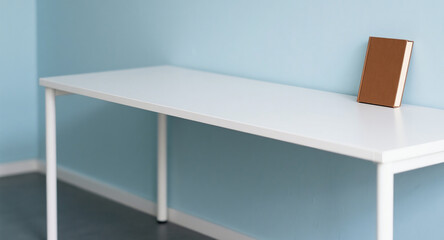 A minimalistic scene with a white table against a light blue wall, featuring a single brown book standing upright on the tabletop.