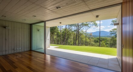Fototapeta premium A modern concrete and wood interior opens to a lush green lawn and distant mountains through a large glass sliding door