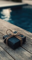 A small, dark gift box tied with twine rests on weathered wooden planks beside a blurred, dark blue swimming pool