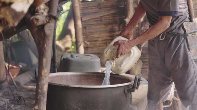 farmer in rural Flores, Indonesia, pouring fermented sap from sugar palm, Arenga pinnata, into metal pot over firewood at a traditional village distillery to produce arak or sopi liquor