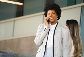 Businesswoman smiling while talking on smartphone outside office building with colleague in...