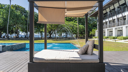 Recreation area by the resort swimming pool. Jets are beating over aquamarine water. A bed with a mattress and pillows under a canopy in the foreground. Green trees in the distance. Philippines. 
