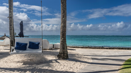 A swing bench is suspended between the trunks of palm trees on a sandy beach. Cushions on the seat. Wicker plastic chairs on the embankment. Turquoise ocean, blue sky, clouds. Resort. Hotel. Mauritius