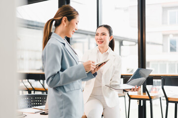 Obraz premium Two Asian businesswomen in modern office, one holding tablet and other with laptop, smiling and discussing work, bright natural light, professional atmosphere