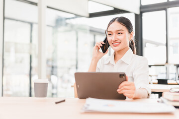 Obraz premium Young woman business attire sitting at desk holding tablet talking on smartphone smiling modern office natural light professional confident productive