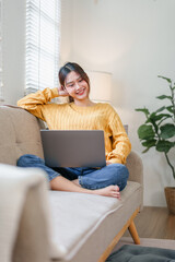 Young woman in yellow sweater sitting on sofa with laptop, smiling and relaxing in cozy living room...