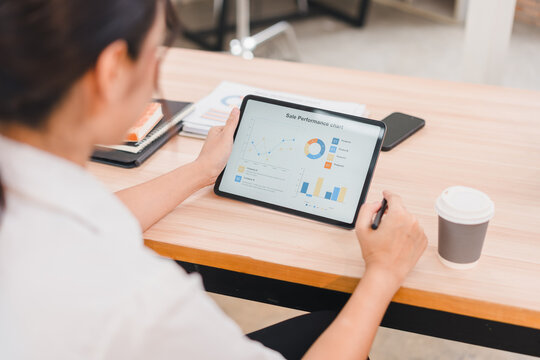 Businesswoman analyzing sales performance chart on digital tablet, reviewing data and graphs at modern office desk with coffee cup, focused and professional atmosphere