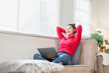 Young woman red sweater blue jeans sitting on sofa relaxing with laptop natural light cozy home...