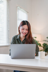 Young Asian woman working on laptop at home office, natural light from window, relaxed atmosphere,...