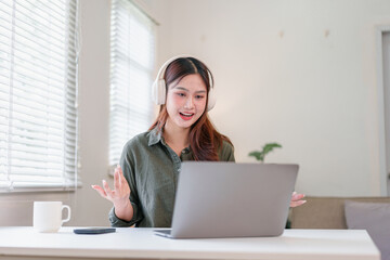 Young Asian woman wearing headphones sits at white desk, smiling and gesturing while video calling...