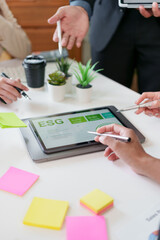 Business team discussing ESG strategy with digital tablet, colorful sticky notes, and potted plants on white office desk, showing collaboration and sustainability focus