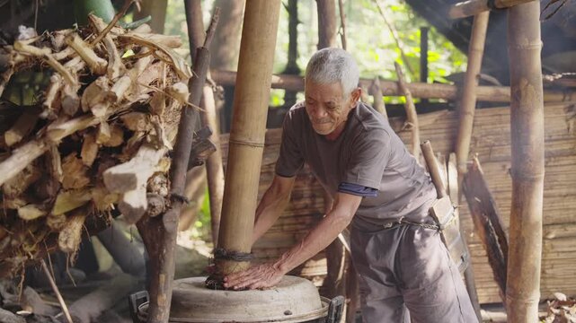 Elderly palm wine farmer working in a rural distillery in Flores, Indonesia, sealing a metal cooking pot with bamboo pipe to distill fermented sap into traditional arak or sopi alcohol