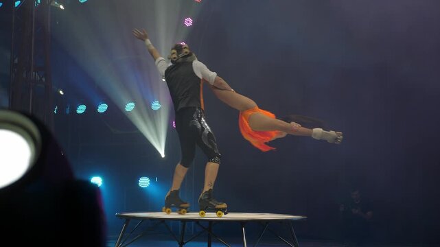 A young couple of acrobats on roller skates perform in a circus. A couple of acrobats perform on roller skates, they rotate around themselves on a small round stage. Concert lighting.