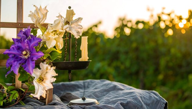 A charming still life arrangement of vibrant purple and cream clematis flowers, illuminated by the soft glow of the setting sun, placed on a textured grey cloth surface near a dark metal candle...