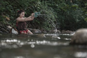 Obraz premium Young Asian woman is happily washing clothes in a natural river, splashing water with a smile. Traditional rural lifestyle scene with green forest background, fresh nature, 