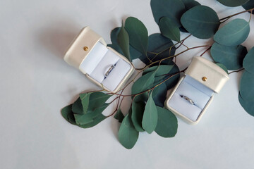 The wedding rings are in boxes on a corner of a white table, decorated with dried flowers