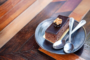 a pice of chocolate cake serving on dessert dish with couple of spoon on wooden table