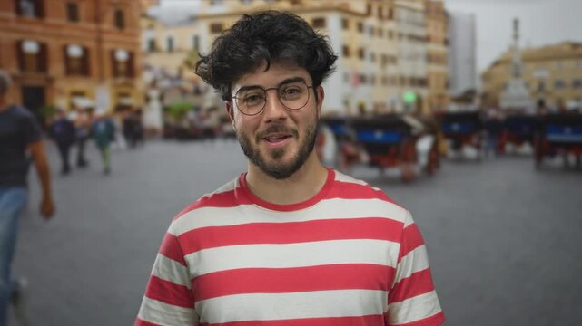 Young hispanic man with beard and glasses smiling on a bustling street outdoors wearing red striped shirt with buildings and people in the background.