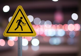Yellow triangular warning sign with a person holding a bright light or tool, sharply defined against a soft, blurred bokeh background of urban lights, emphasizing caution