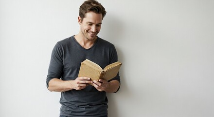 A smiling man in a gray shirt reads an open book against a white wall