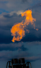A powerful burst of vibrant orange flame erupts from a metal burner. The complex structure of the burner is visible beneath the fire, with coils and pipes suggesting the immense heat generated.