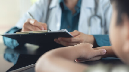 Female pediatrician doctor and a young boy patient at clinic office. Medical work writing a prescription on a table in a hospital.