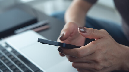 Close-up of woman using smartphone while working on laptop computer at home office, working remotely, freelance at work, online job, brown tone