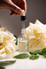 A hand carefully extracts rose oil from a dropper, surrounded by fresh white roses and green...
