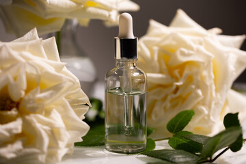 A glass vial filled with oil stands next to delicate rose buds. Surrounded by soft petals and green...