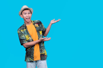 Excited Indonesian Man In A Hawaiian Shirt And Hat, Presenting With Open Palms On A Blue Background