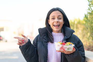 Young woman at outdoors holding a bowl of fruit at outdoors surprised and pointing finger to the...