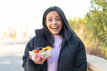 Young woman at outdoors holding a bowl of fruit at outdoors with surprise and shocked facial...