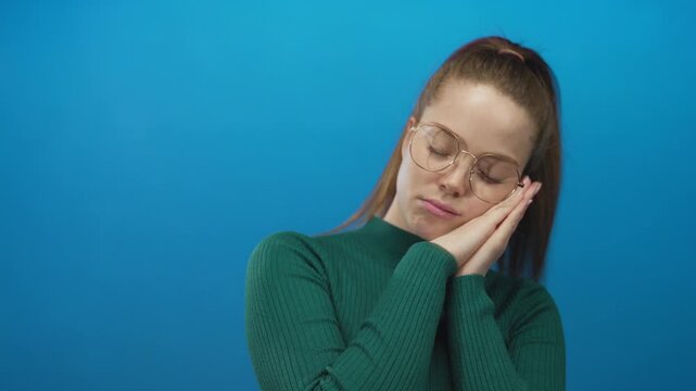 Young caucasian woman in green sweater with glasses pressing clasped hands to cheek against blue wall in studio; sleepiness.