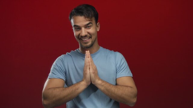 Young hispanic man smiling with hands in namaste gesture wearing blue shirt against isolated red background expressing gratitude and serenity